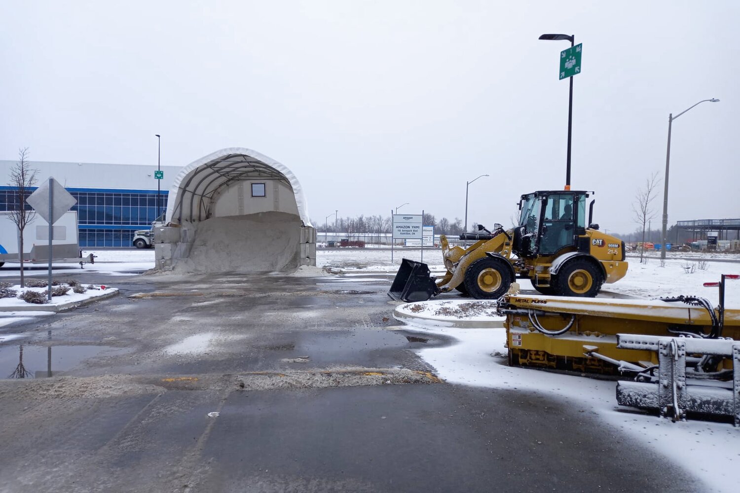 Yellow CAT loader and de-icing equipment on a wet, snowy industrial parking area beside a covered salt pile.
